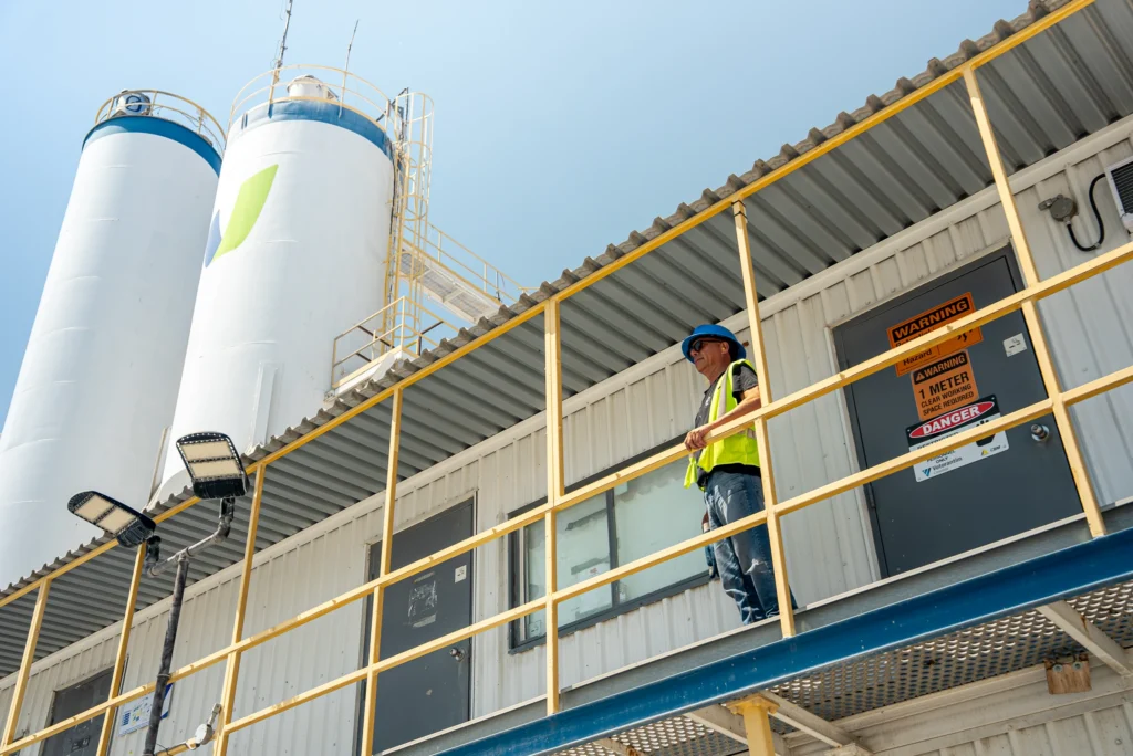 a worker in PPE standing on the second level of a concrete plant building looking into the distance with plant silos in the background.